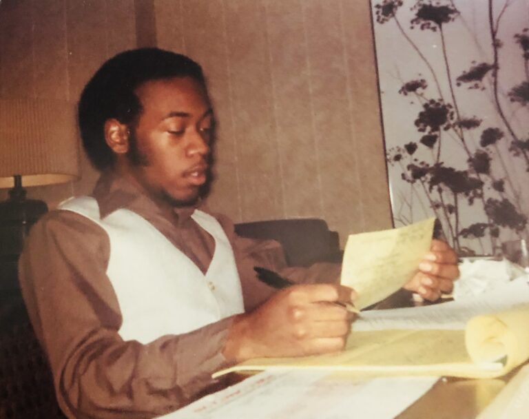 A young Danté sits at a desk, reviewing notes from a yellow legal pad. He is wearing a brown shirt and white vest.